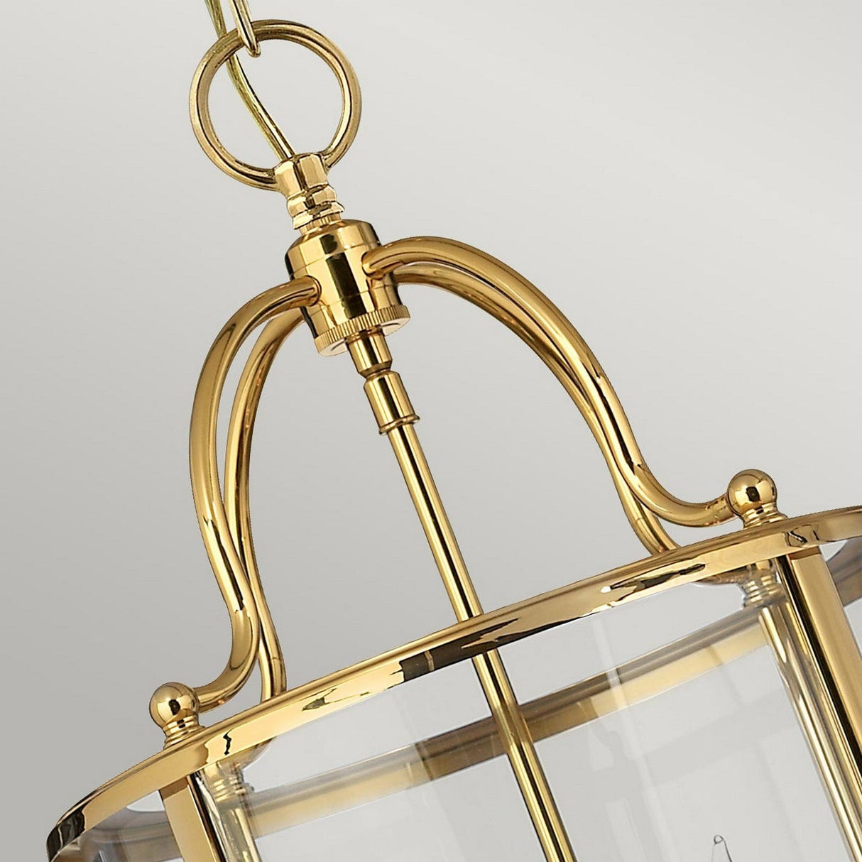 Close-up of the Gentry 4 Light Medium Pendant in polished brass, showcasing its clear bent glass panels. The traditional design features elegant curves and a circular frame, suspended by a chain against a light, blurred background.
