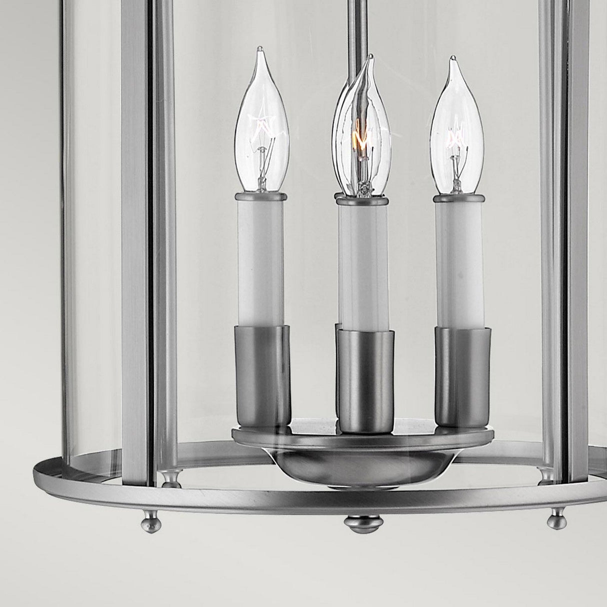A close-up of the Gentry 4 Light Medium Pendant - Silver shows a brushed nickel chandelier featuring three candle-shaped light bulbs, enclosed within clear bent glass panels. The design presents a modern, sleek appearance.
