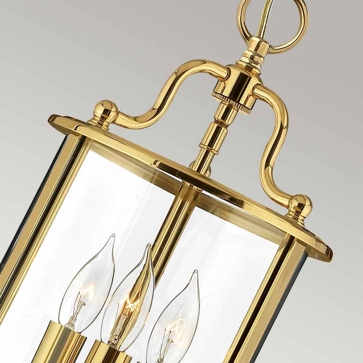 A close-up of the Gentry 3 Light Small Pendant in polished brass showcases its clear glass casing. Inside, three candle-shaped bulbs emit a warm glow. The fixture's polished finish and classic design offer an elegant vintage look.