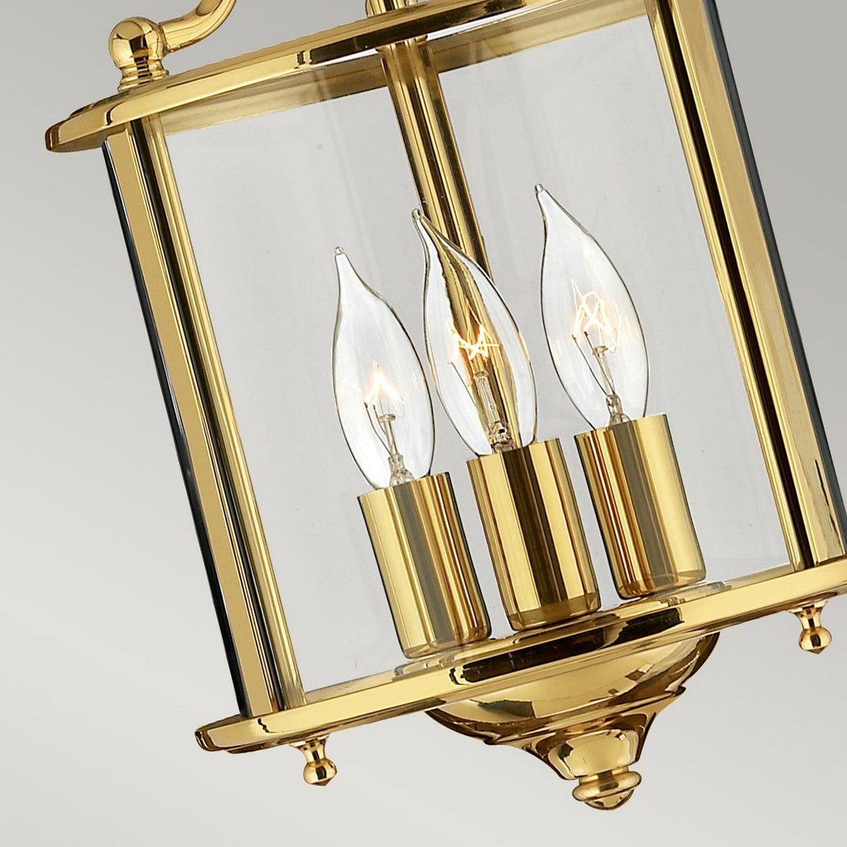 Close-up of the Gentry 3 Light Small Pendant in polished brass, featuring three candle-shaped bulbs enclosed in clear glass panels, showcasing a traditional design against a gray background.