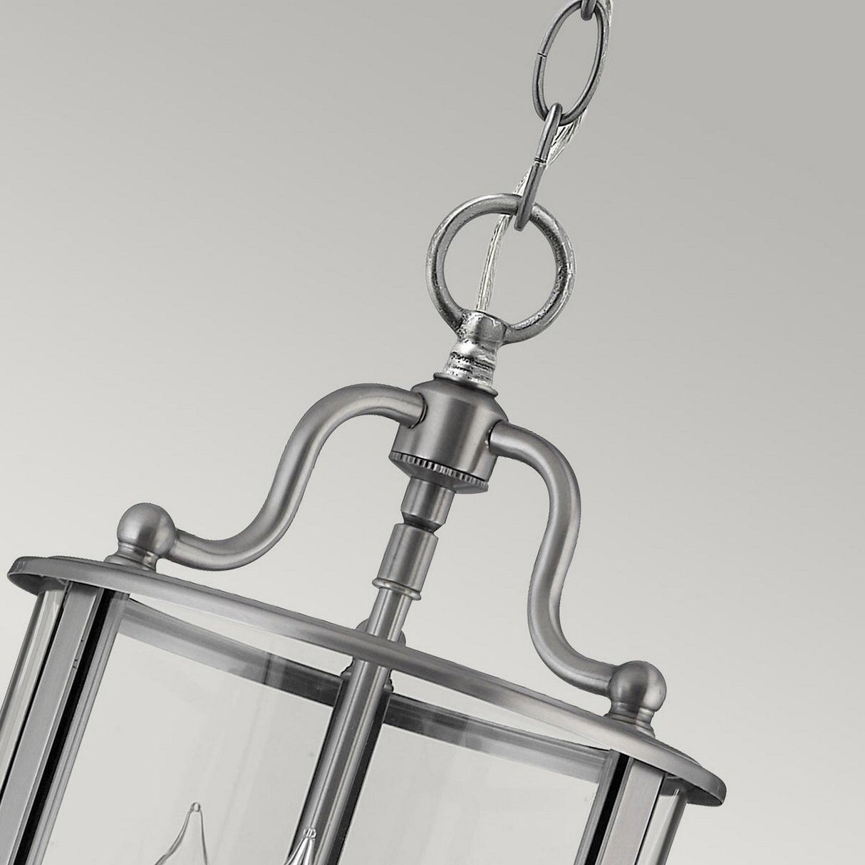 A close-up view of a hanging lantern-style light fixture showcases a metallic frame with clear bent glass panels, suspended by a decorative chain in a silver finish, set against a gray background. This design is similar to the Gentry 3 Light Small Pendant - Silver.