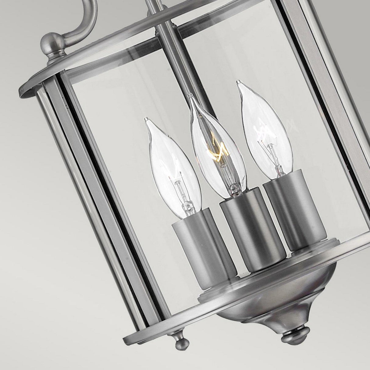 Close-up of a modern silver finish lantern-style light fixture featuring three candle-shaped bulbs inside, encased in clear bent glass panels against a neutral gray background, exemplifying the elegance of the Gentry 3 Light Small Pendant - Silver.