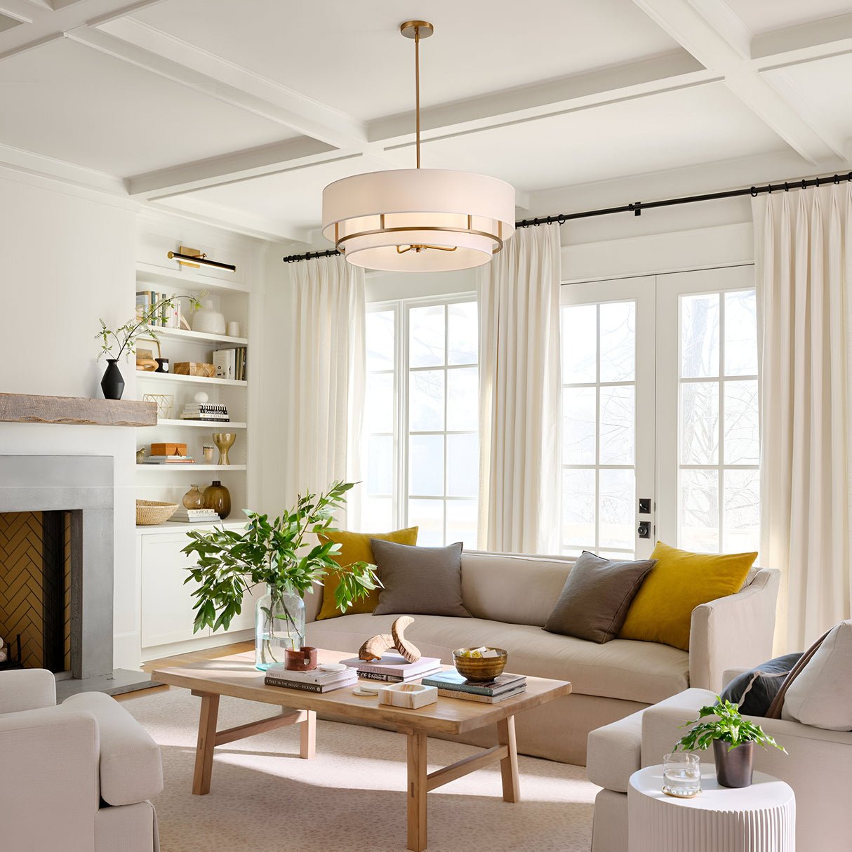 A modern living room features a beige sofa and armchair, adorned with yellow and gray cushions. A wooden coffee table with books and plants sits below a Graham 4 Light Duo-Mount Pendant/Semi-Flush in lacquered brass, elegantly hanging from the coffered ceiling. Built-in shelves, a fireplace, and white curtains complete the scene.