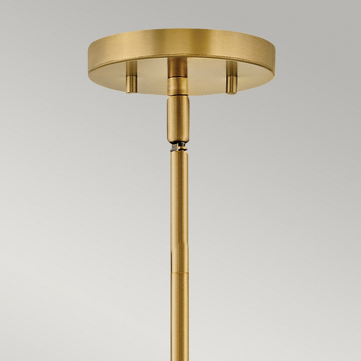 Close-up of a modern ceiling light fixture featuring a round brass canopy and connecting rod, highlighting the dual-mount capability of the Graham 3 Light Duo-Mount Pendant/Semi-Flush in Lacquered Brass. The fixture stands out against a plain, light gray background.