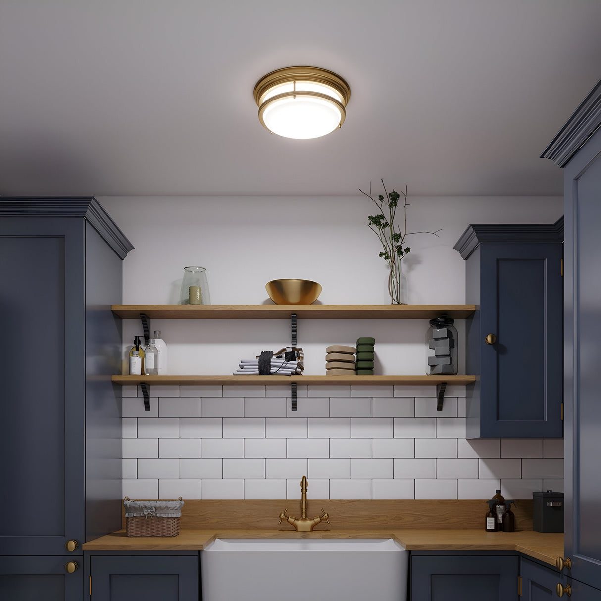A kitchen featuring navy cabinets, white subway tiles, and wooden countertops radiates charm. Two wooden shelves display ceramic dishes, jars, and a vase with branches. A Hadley 3 Light Flush - Brushed Bronze fixture illuminates the space above a farmhouse sink complemented by a brushed bronze faucet.