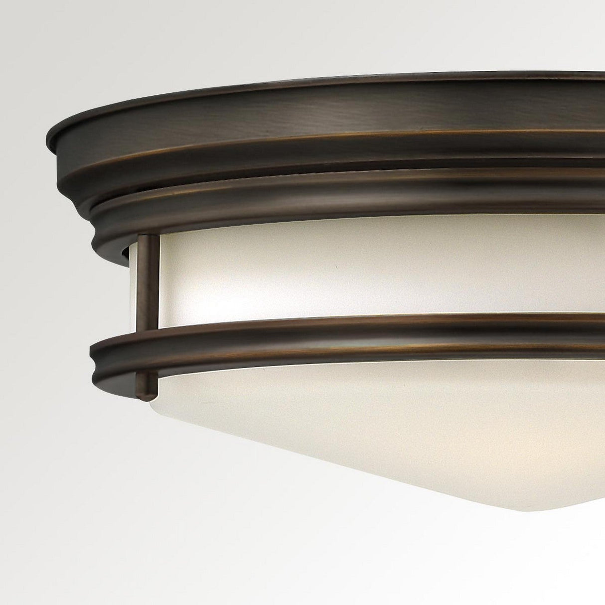 A close-up view of the Hadley 3 Light Flush in bronze showcases its retro design with a frosted glass shade. The fixture features modern clean lines and circular elements, mounted seamlessly against a plain white ceiling.