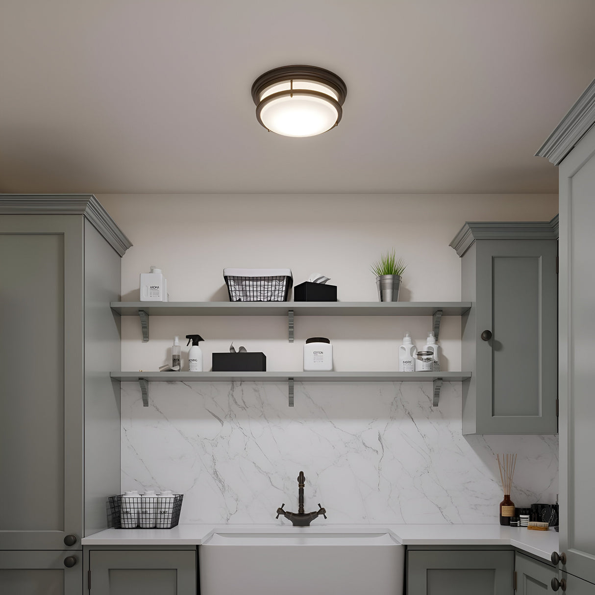 The laundry room boasts gray cabinets and shelves adorned with a white marble backsplash. The shelves neatly organize baskets, cleaning products, and a potted plant. The space is illuminated by the Hadley 3 Light Flush in Bronze, casting light over a deep sink fitted with a bronze faucet.