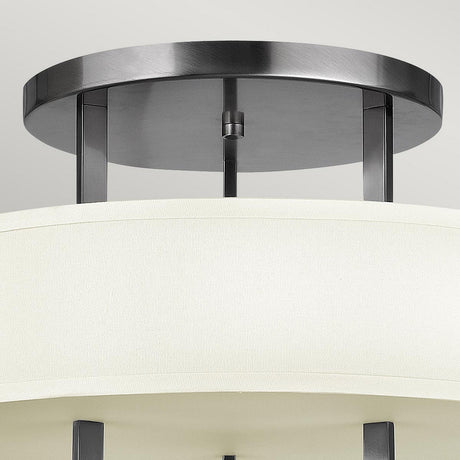 Close-up of a Hampton 3 Light Semi Flush ceiling fixture, featuring a circular metal base and an ivory drum shade. The elegant metal supports in an Antique Nickel finish enhance its contemporary appearance.