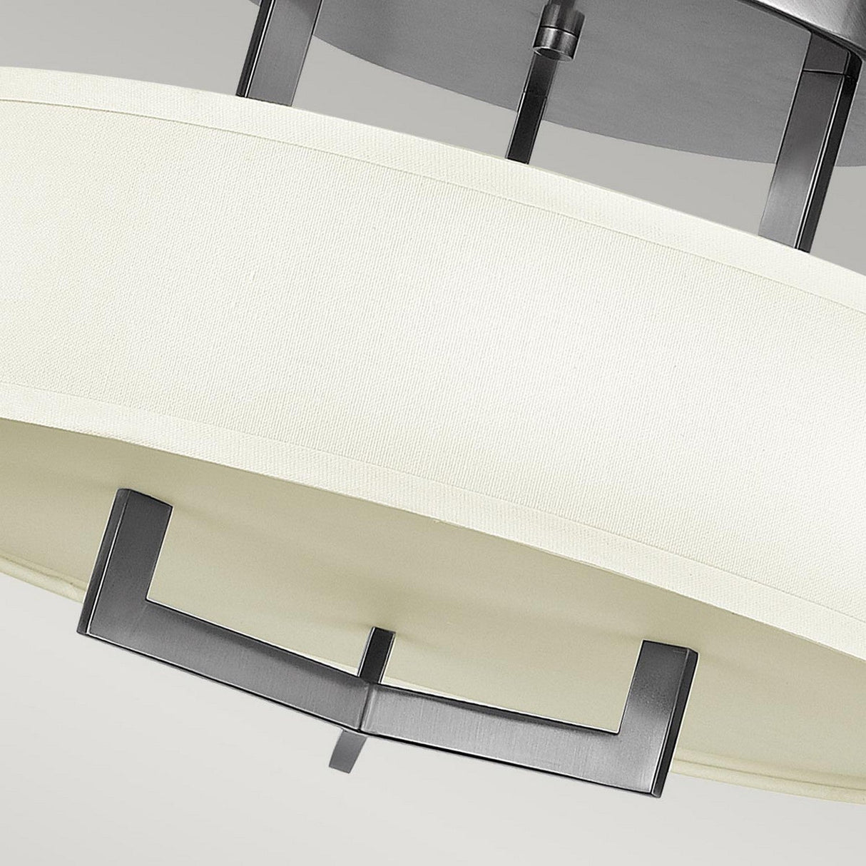 A close-up of the Hampton 3 Light Semi Flush reveals its modern ceiling light design, featuring ivory linen drum shades and sleek metal accents in Antique Nickel. This contemporary fixture showcases clean metallic lines against a neutral backdrop.