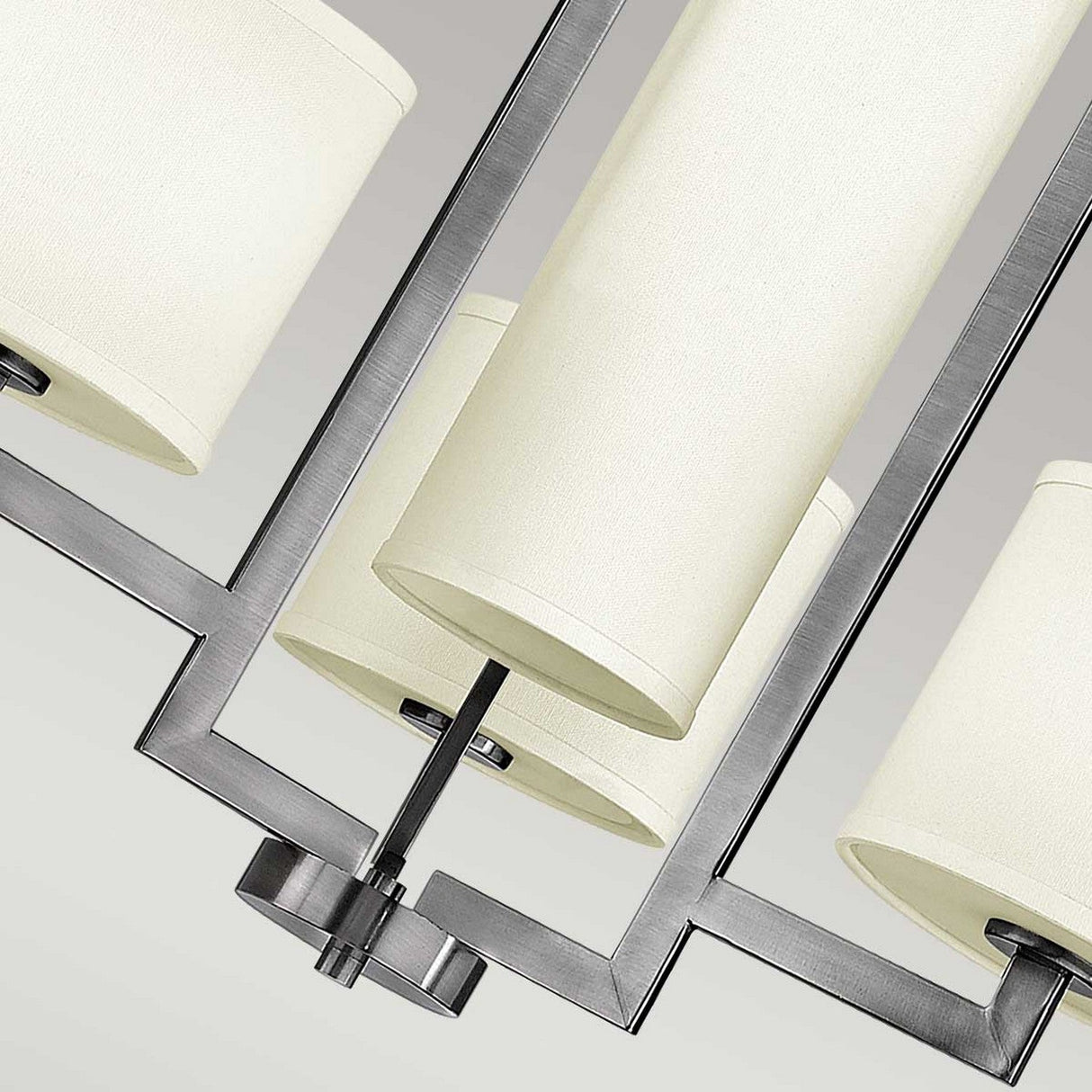 Close-up of a Hampton 4 Light Chandelier in transitional design, showcasing an antique nickel finish with cylindrical ivory lampshades and a geometric metal frame, viewed from below against a neutral background.