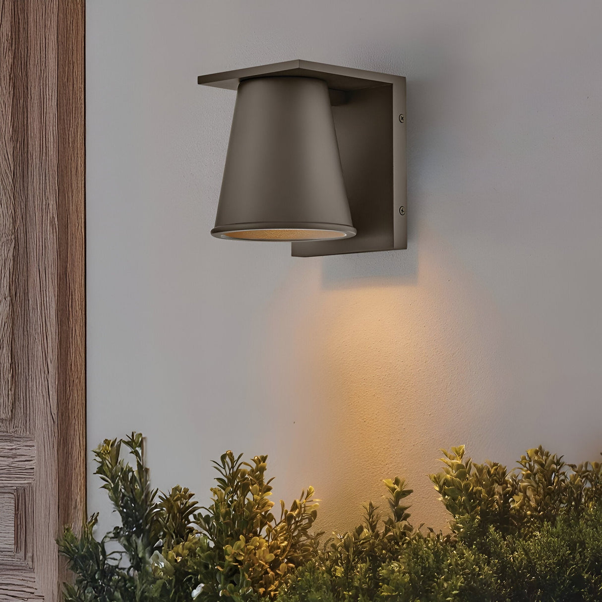 The Coastal 1 Light Small Outdoor Wall Lantern in architectural bronze casts a warm glow on a white wall above green shrubs, next to a wooden door frame. Weather-resistant and modern, it’s ideal for outdoor spaces.