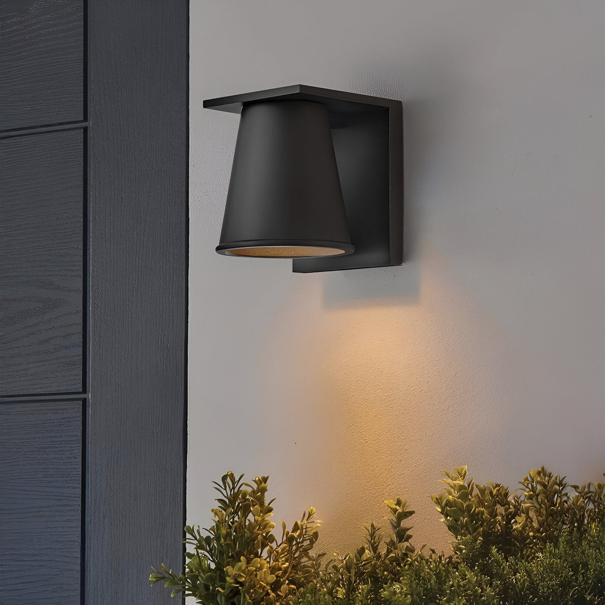 The Coastal 1 Light Small Outdoor Wall Lantern in textured black features a minimalist design with clean lines. Mounted on a white wall, it casts warm light downward onto green shrubs, ideal for modern outdoor lighting.