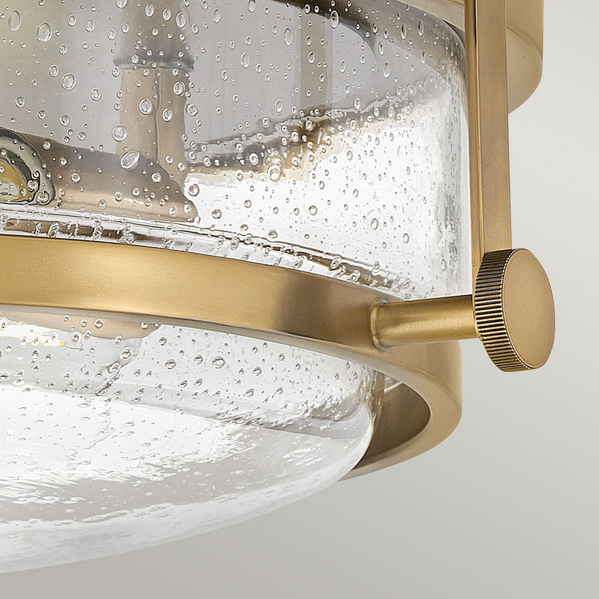 Close-up of the Harper 3 Light Flush fixture in Heritage Brass, featuring a cylindrical clear seeded glass cover. The bubbled texture of the glass and contemporary design incorporate retro elements with rounded edges and visible fasteners, creating a seamless flush mount look.