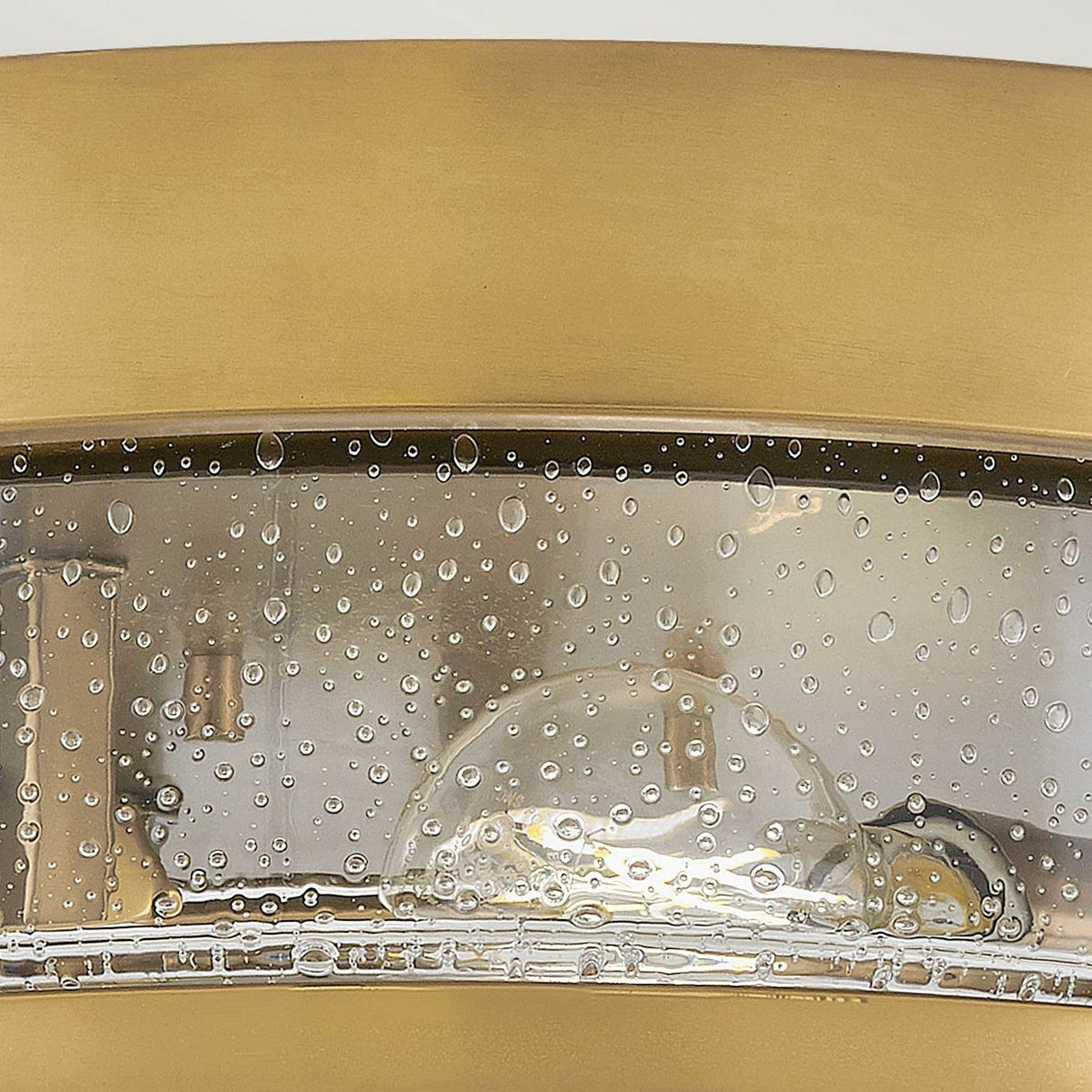 Close-up of the Harper 3 Light Flush in Heritage Brass with Clear Seeded Glass. The retro design features glass speckled with water droplets, highlighting the shiny surface and detailed craftsmanship underneath, reminiscent of a classic flush mount fixture.