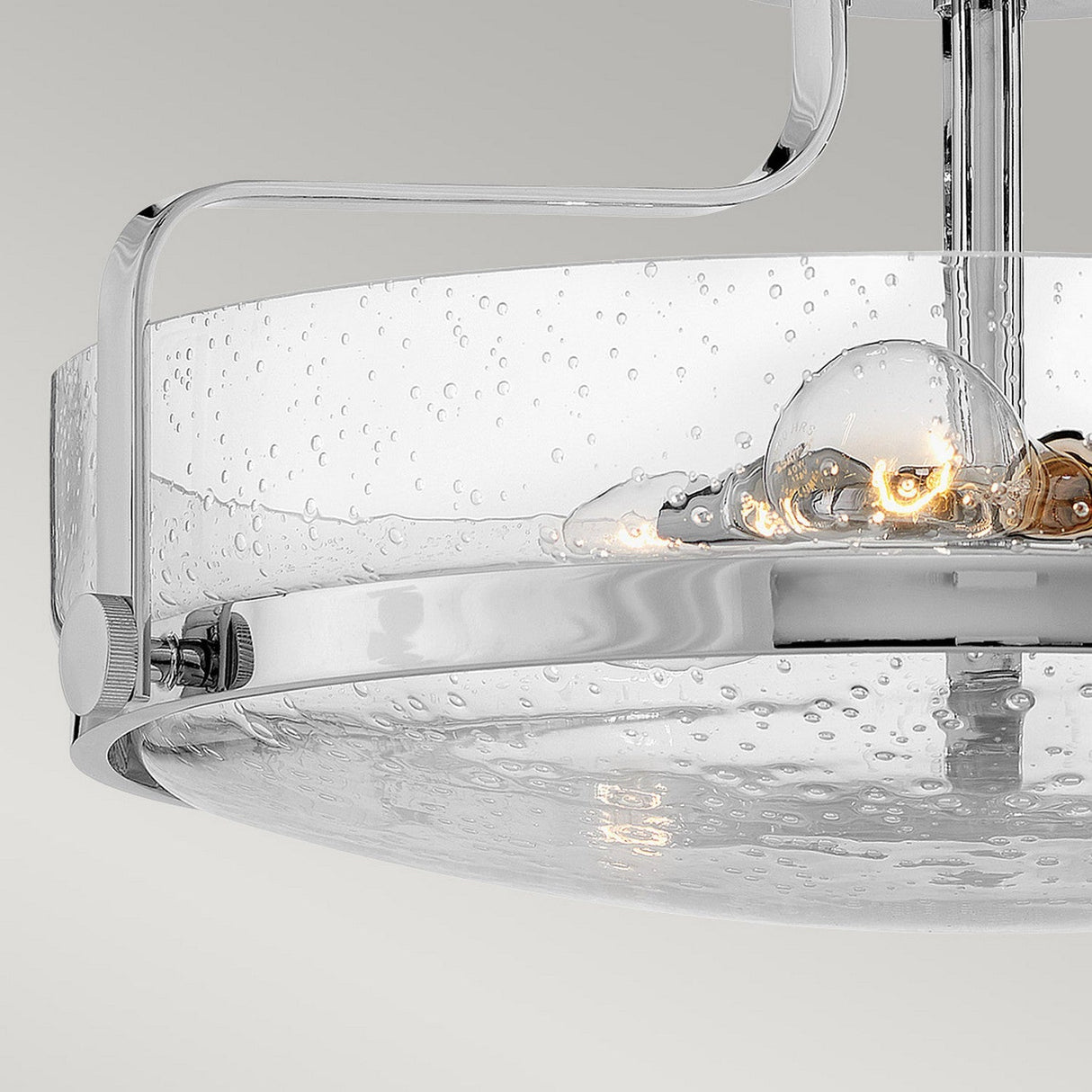 Close-up of the Harper 3 Light Medium Semi Flush, showcasing its contemporary design with a circular clear seeded glass shade. Inside, a visible bulb emits a warm glow. The fixture is accented by a sleek polished chrome frame, set against a neutral gray background.