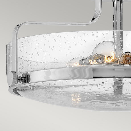 Close-up of the Harper 3 Light Medium Semi Flush, showcasing its contemporary design with a circular clear seeded glass shade. Inside, a visible bulb emits a warm glow. The fixture is accented by a sleek polished chrome frame, set against a neutral gray background.
