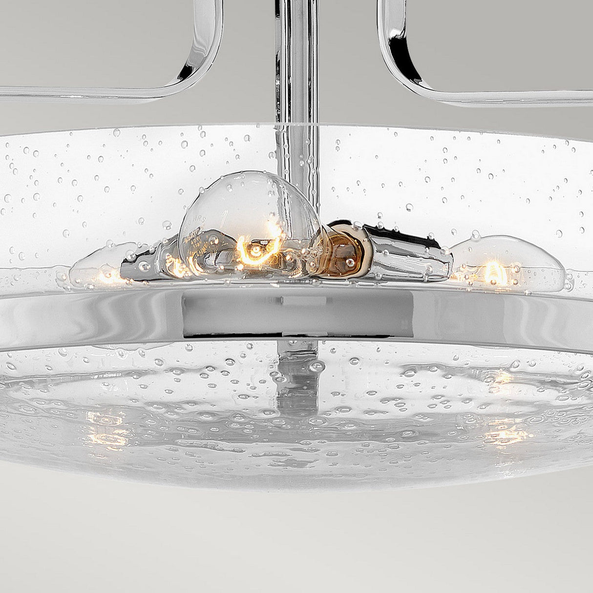 A close-up of the Harper 3 Light Medium Semi Flush showcases its contemporary design, featuring a polished chrome finish and a clear seeded glass cover. The glowing light bulbs within highlight its elegant components against a neutral backdrop.