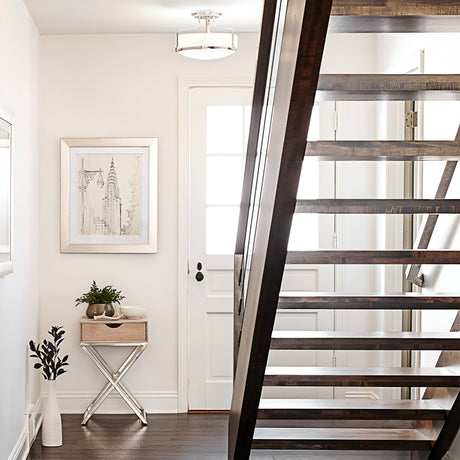 A modern hallway features a wooden staircase and a white door with glass panels. The space is illuminated by the Harper 3 Light Medium Semi Flush in Polished Chrome with Opal Etched Glass. Below a framed sketch of the Chrysler Building, a small side table with a plant enhances the minimalist decor and neutral tones.