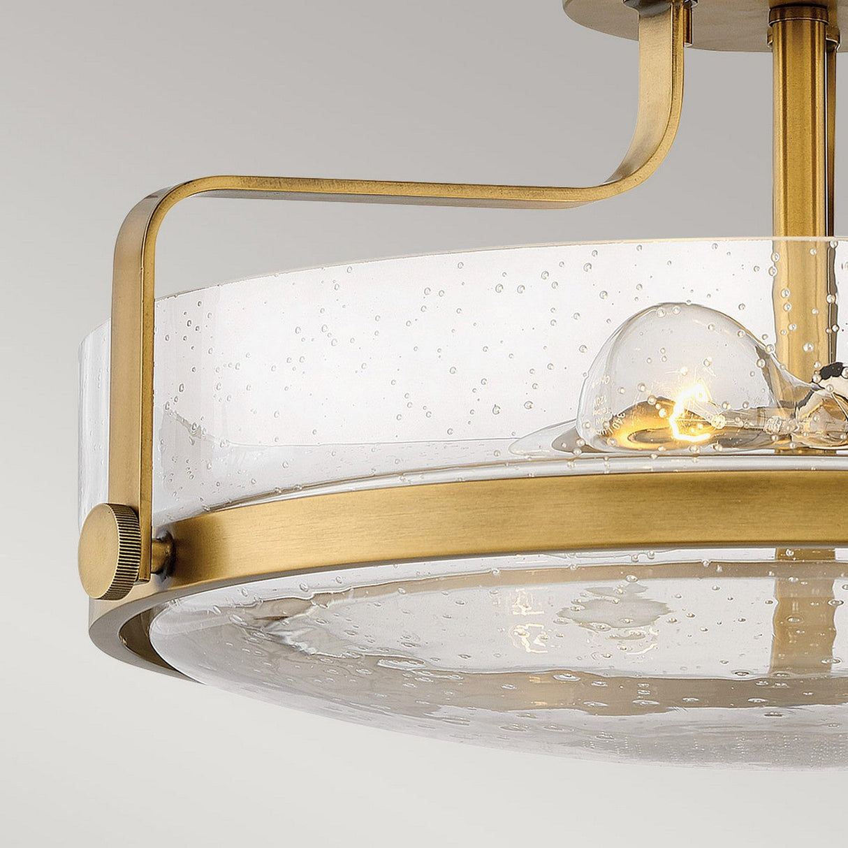 The Harper 3 Light Medium Semi Flush in Heritage Brass features a contemporary design with a frame and a clear seeded glass shade adorned with small bubbles. The visible bulb inside provides a warm glow, enhancing the ambiance beautifully.