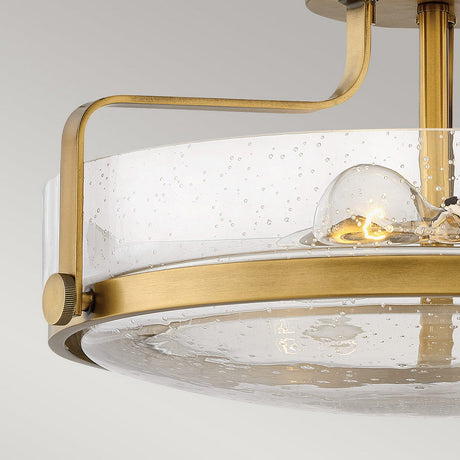 The Harper 3 Light Medium Semi Flush in Heritage Brass features a contemporary design with a frame and a clear seeded glass shade adorned with small bubbles. The visible bulb inside provides a warm glow, enhancing the ambiance beautifully.