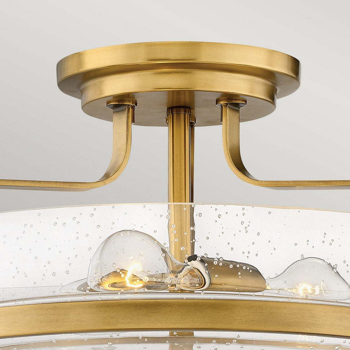 The Harper 3 Light Medium Semi Flush - Heritage Brass with Clear Seeded Glass features a detailed close-up of a semi-flush mount ceiling light fixture. It showcases three exposed bulbs within a clear seeded glass shade, adorned with a bubble pattern, while its heritage brass finish enhances the modern design, complete with a round base and elegantly curved supports.
