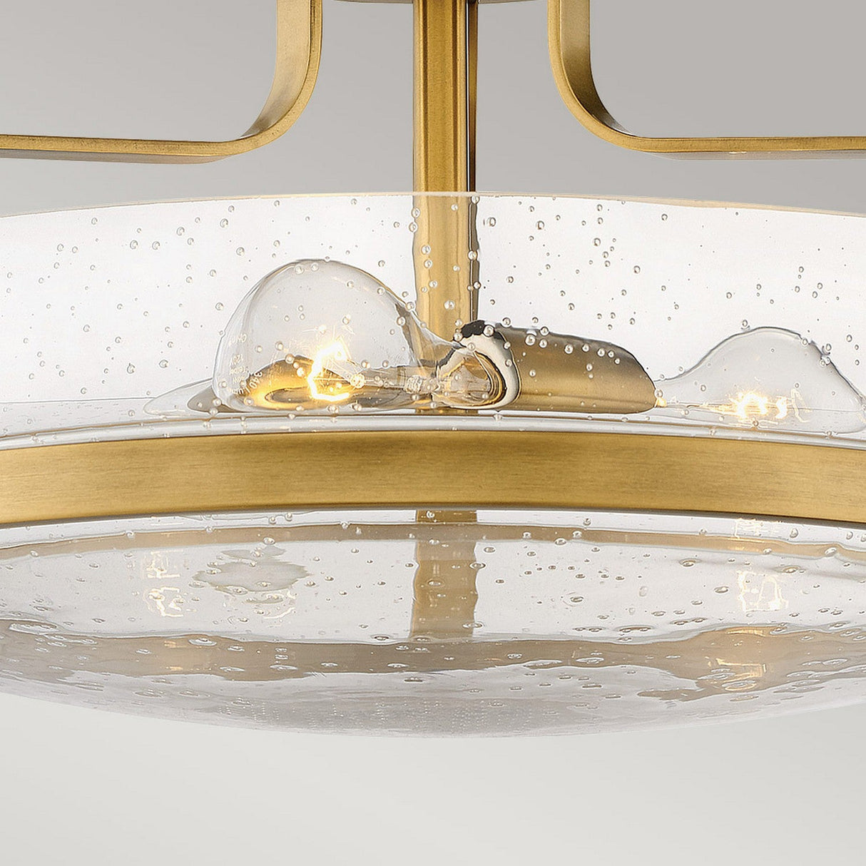 A close-up view showcases the Harper 3 Light Medium Semi Flush, featuring a circular design in heritage brass. This modern chandelier includes a clear seeded glass dome surrounding two visible light bulbs, creating a bubble-like appearance against a neutral background.