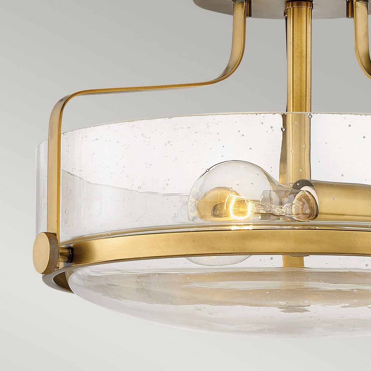 The Harper 3 Light Small Semi Flush in Heritage Brass with Clear Seeded Glass showcases a contemporary design, featuring clear seeded glass and heritage brass accents. This semi-flush mount fixture reveals a visible incandescent bulb, while its brass arms elegantly curve to support the design against a plain light gray background.