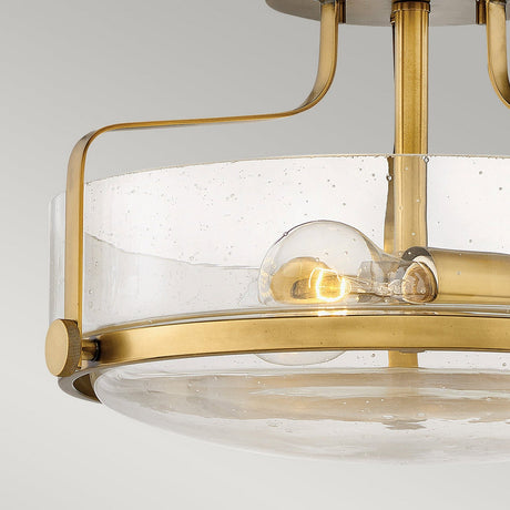 The Harper 3 Light Small Semi Flush in Heritage Brass with Clear Seeded Glass showcases a contemporary design, featuring clear seeded glass and heritage brass accents. This semi-flush mount fixture reveals a visible incandescent bulb, while its brass arms elegantly curve to support the design against a plain light gray background.