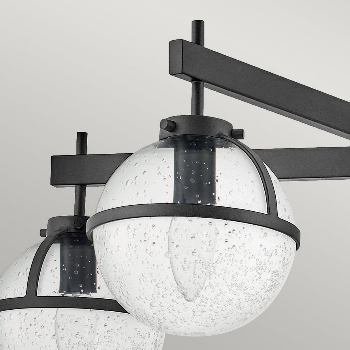A close-up of the Hollis 5 Light Semi Flush showcases its mid-century modern design, featuring two spherical clear seeded glass shades filled with tiny bubbles, each elegantly encased in sleek black metal frames. This minimalist black semi-flush light includes a horizontal black metal bar that complements its aesthetic.
