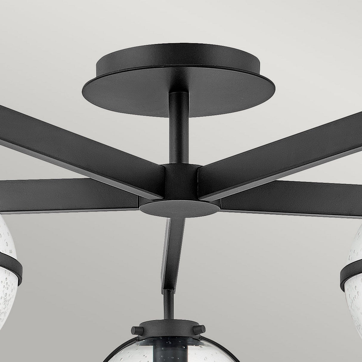 A detailed view of the Hollis 5 Light Semi Flush in black showcases its five arms radiating outward. Each arm supports a clear seeded glass globe with a speckled design, reflecting mid-century modern lighting style, against a simple, light-toned ceiling backdrop.