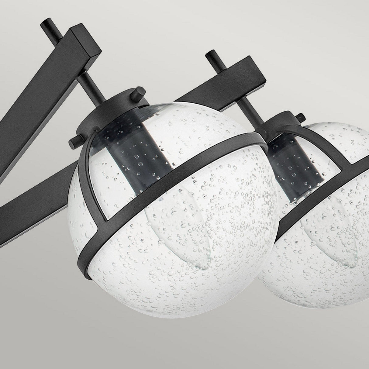 A close-up of the Hollis 5 Light Semi Flush in black, featuring two spherical clear seeded glass shades with bubble-like patterns. Each shade is set within a black metal frame, and the fixture is elegantly mounted on a black bar. This design adds sophisticated flair against the gray background.