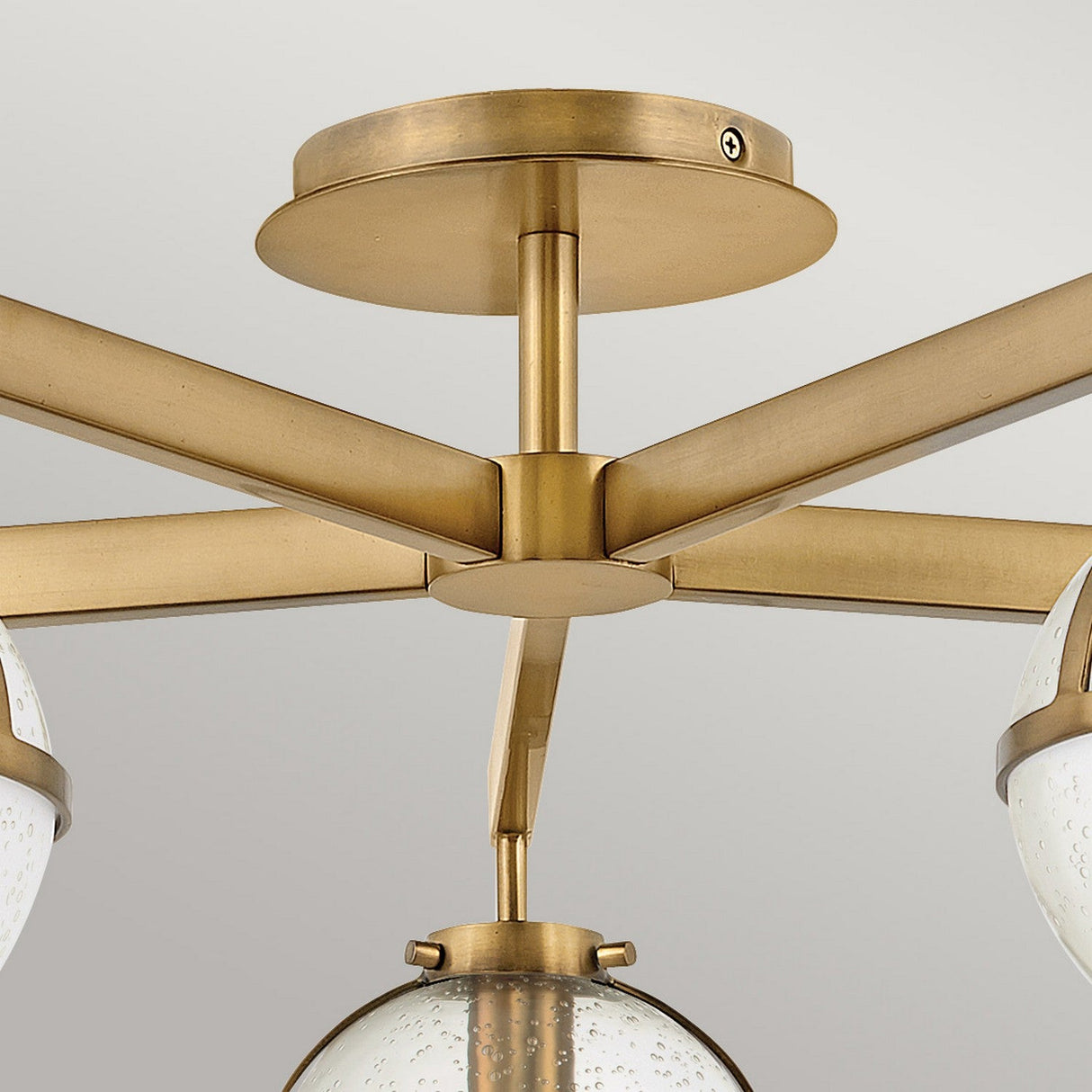Close-up view of the Hollis 5 Light Semi Flush fixture in Heritage Brass, featuring multiple arms that support spherical clear seeded glass shades. The design showcases a sleek, minimalist style against a light gray background.