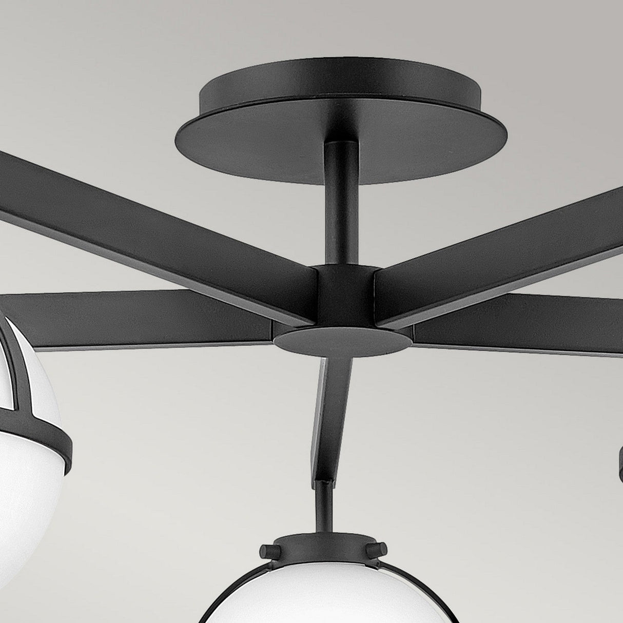 The Hollis 5 Light Semi Flush fixture showcases a contemporary design with round opal etched glass globes. Its sleek, geometric structure includes a black finish, featuring a central mounting plate from which arms extend outward to support the globes. The background is plain gray.