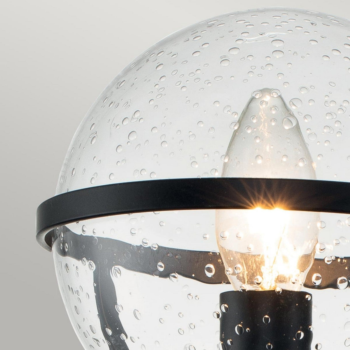 A close-up of the Hollis 1 Light Wall Light showcases its stunning bubble glass globe with a lit bulb inside. The clear glass, perfect for bathroom lighting, is adorned with numerous small bubbles and encircled by a sleek black metal band, adding a modern, Art Deco-inspired elegance.