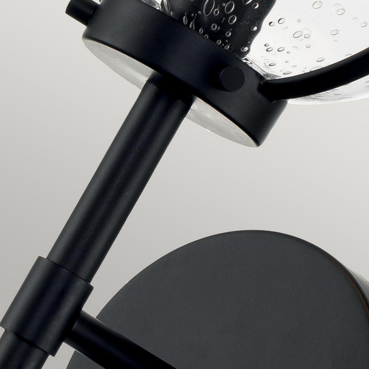 A close-up view features the Hollis 1 Light Wall Light in black with clear glass, highlighting its minimalist and industrial design. Evoking Art Deco lighting, the emphasis is on the lamp's stem and circular components, set against a gray backdrop.