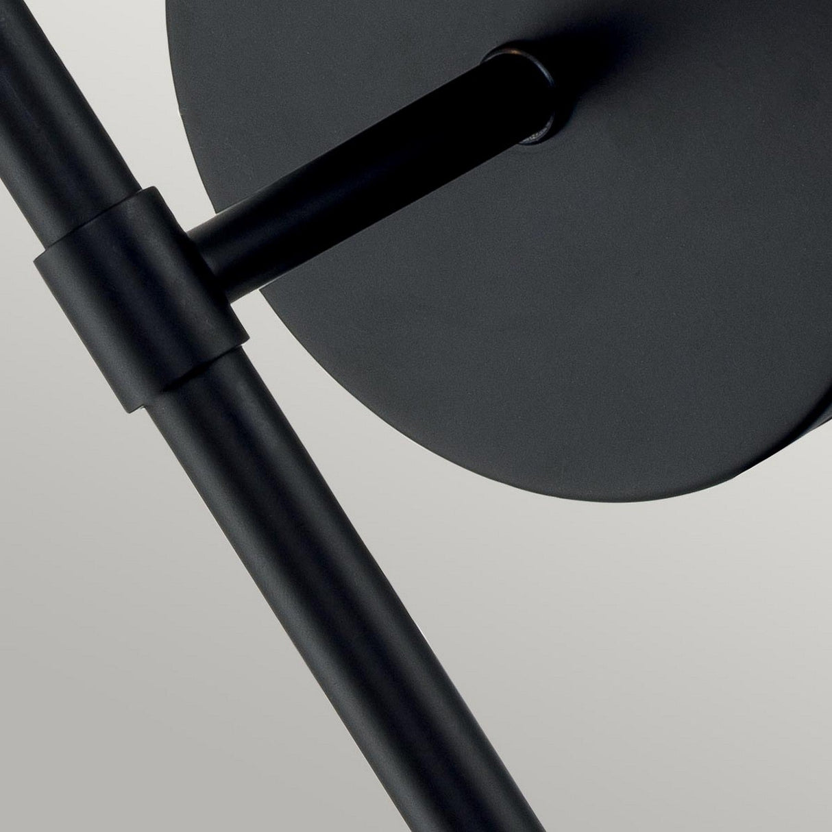 Close-up of the Hollis 1 Light Wall Light in black, featuring a modern metal structure with cylindrical rods intersecting at right angles, reminiscent of Art Deco lighting design. It is attached to a round disk, while the light gray background highlights its sleek elegance and clear glass detailing.