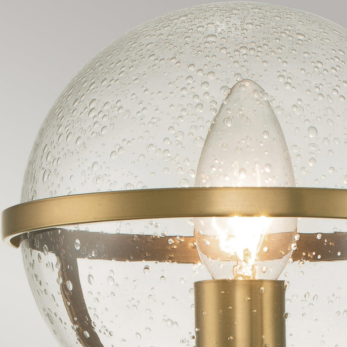 A detailed look at the Hollis 1 Light Wall Light - Heritage Brass With Clear Glass reveals its spherical form with a clear glass shade accented by a bubble pattern. A bulb sits within, framed by a Heritage Brass rim and support, while the textured glass diffuses light to produce an Art Deco-inspired glow.
