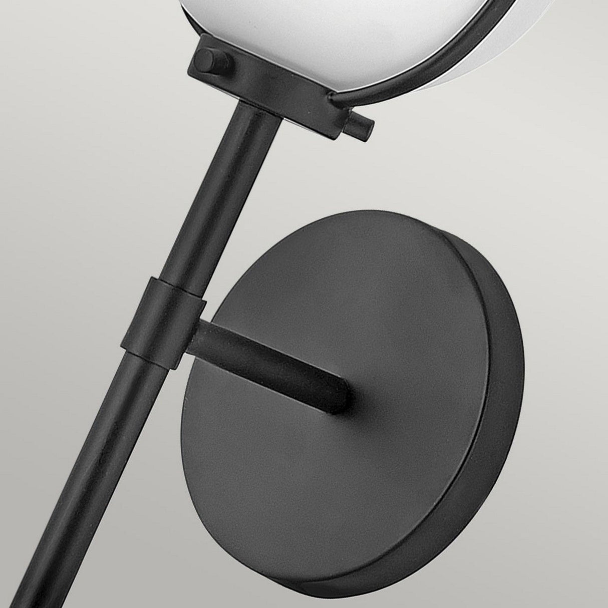 Close-up view of the Hollis 1 Light Wall Light in black, showcasing its modern Art Deco design. It features a circular base and a sleek rod that connects to an opal glass spherical lamp, set against a plain light gray background.