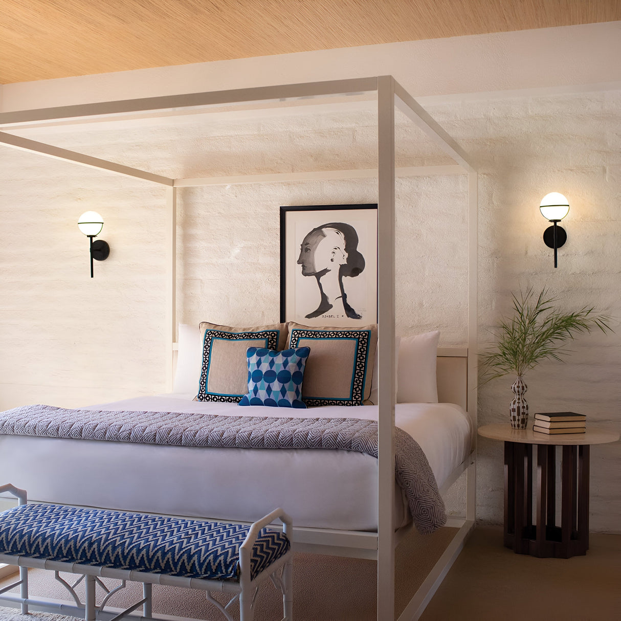 A stylish Art Deco bedroom showcases a white canopy bed decorated with blue and patterned pillows. A framed abstract portrait is displayed on a textured wall, illuminated by the soft light of the Hollis 1 Light Wall Light in black with opal glass. Beside the bed sits a small round table topped with a vase of leaves.