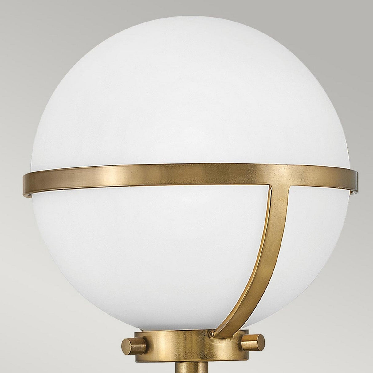 The Hollis 1 Light Wall Light showcases a spherical opal glass lamp encased in a heritage brass frame, highlighted by a thin band encircling the globe and two elegantly curved supports affixed to the base. The plain light gray background complements its Art Deco style.