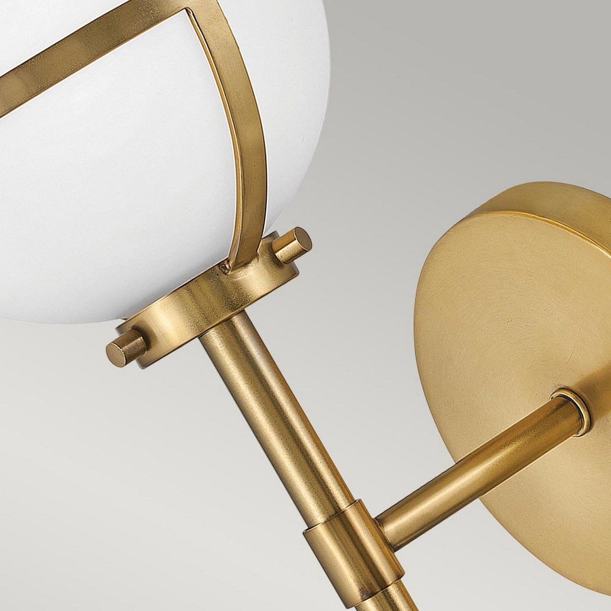 Close-up of the Hollis 1 Light Wall Light, showcasing its opal glass shade paired with heritage brass fixtures. The design includes a sleek cylindrical arm that connects the shade to a circular brass wall mount, evoking the elegance of Art Deco style. The background is a subtle gradient of gray.