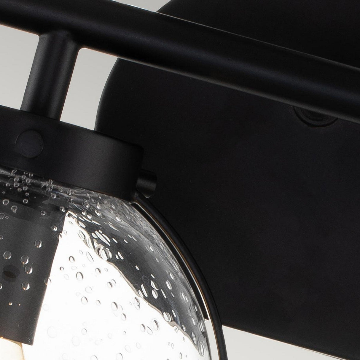 Close-up of the Hollis 2 Light Wall Light, showcasing its sleek black metal design. It includes a clear glass globe adorned with textured water droplet patterns that encases a glowing bulb, offering a modern and elegant appearance.