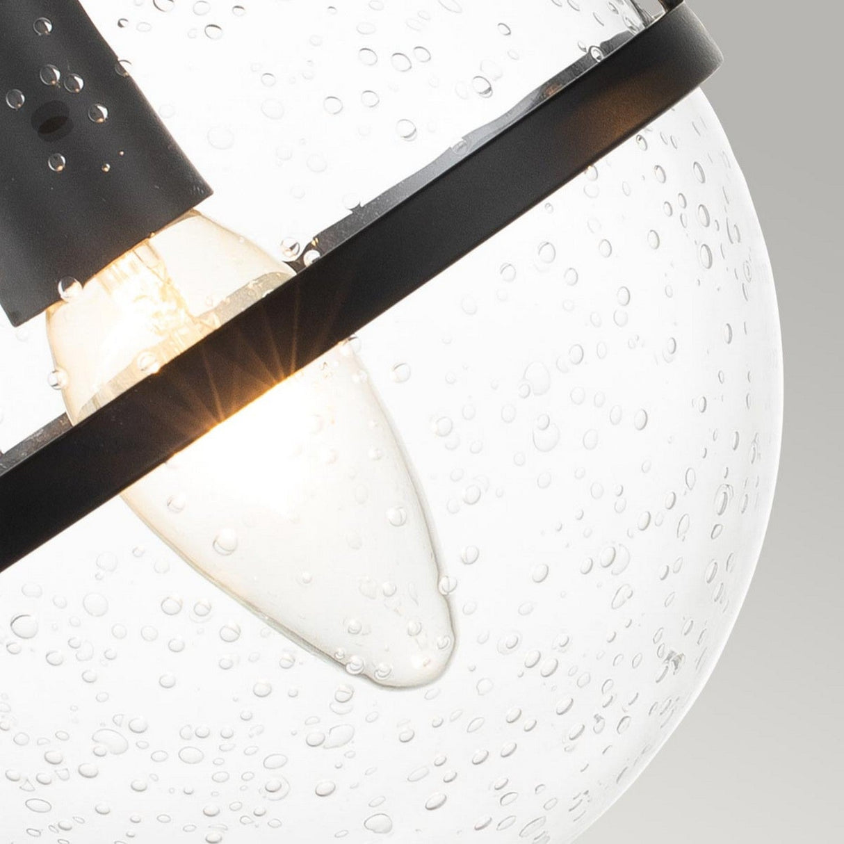 Close-up of a lit Hollis 2 Light Wall Light - Black With Clear Glass, featuring a round, clear glass fixture with delicate water droplet details. This Art Deco lighting piece is accented by a black finish ring, set against a neutral background.