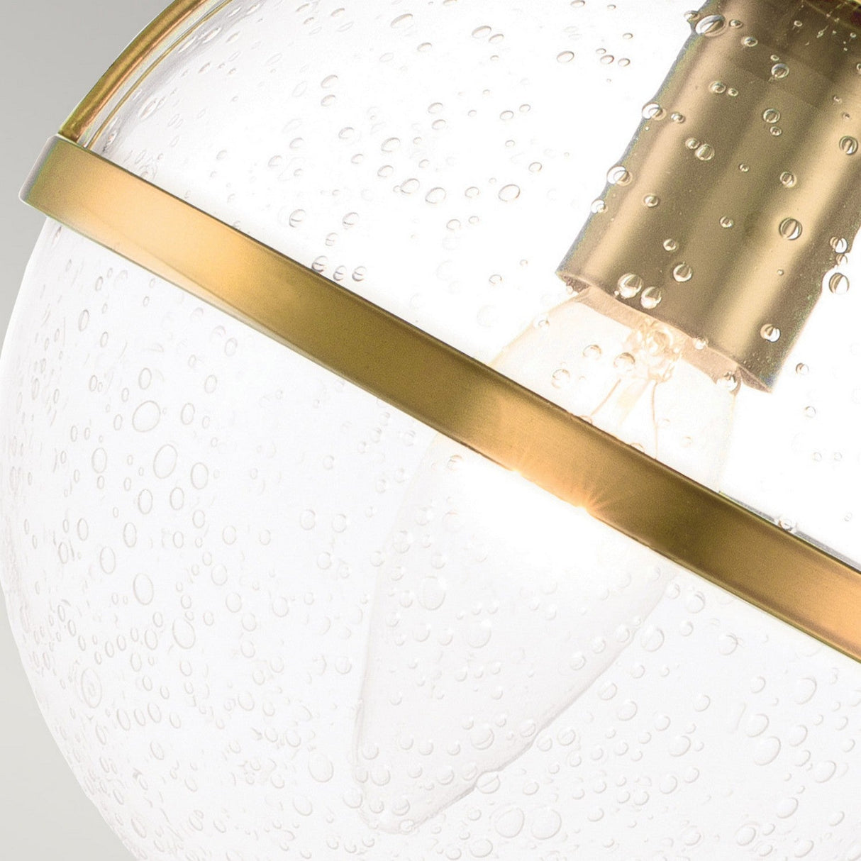 Here is a close-up of the Hollis 2 Light Wall Light, displaying a spherical clear glass design enhanced by a heritage brass band. The bubble-patterned textured glass exposes an inner bulb that emits a warm glow.