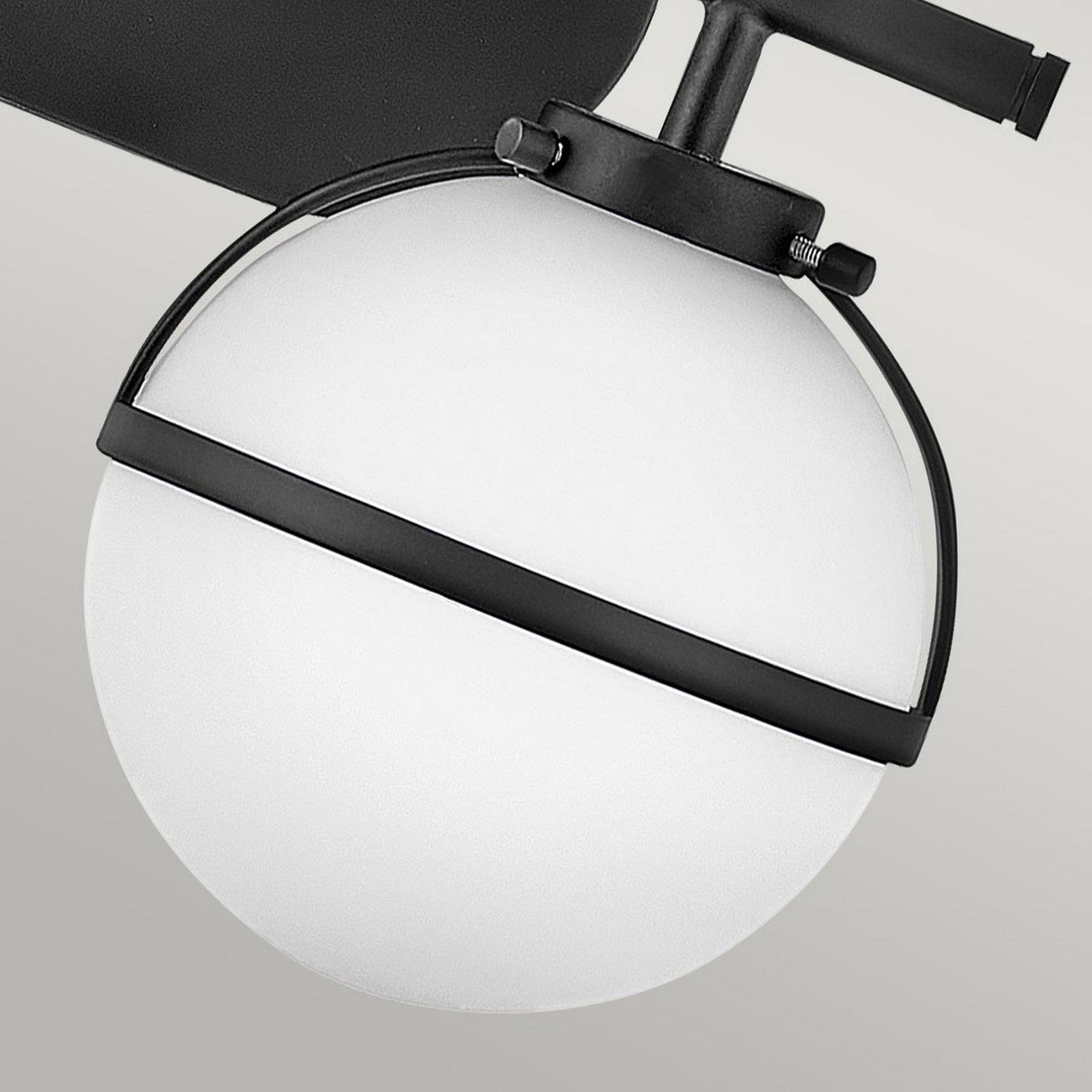 A close-up of the Hollis 2 Light Wall Light reveals its modern, spherical design featuring an opal glass shade accented by a black metal band and handle. The light, with its sleek black finish, is mounted on a grey wall, showcasing a minimalist and contemporary aesthetic.
