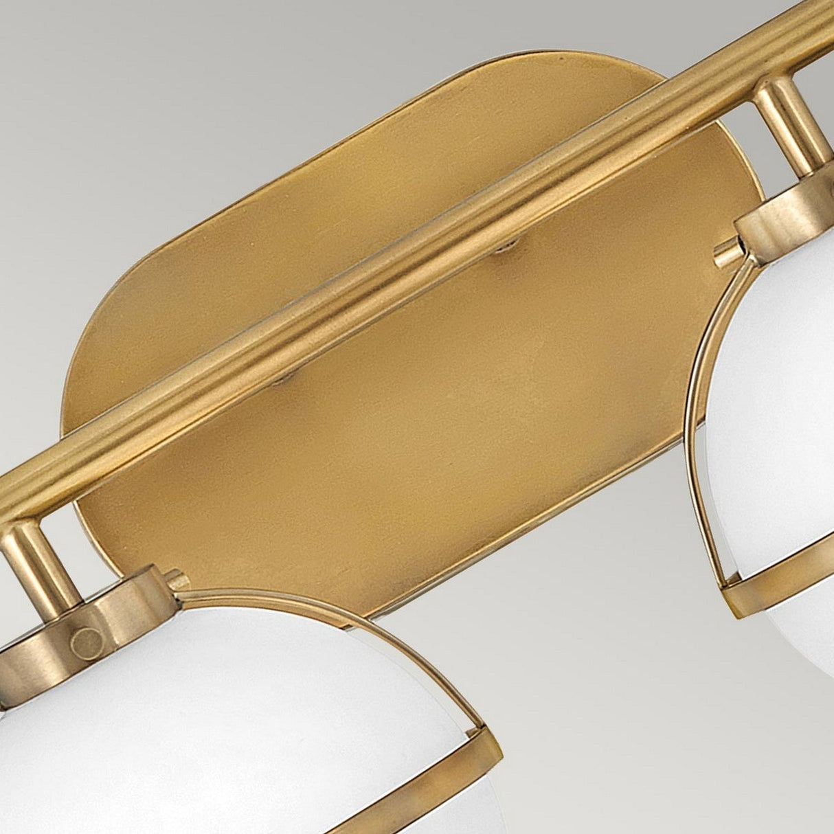 A close-up of the Hollis 2 Light Wall Light in Heritage Brass showcases its two opal glass globes. This fixture features a modern design with a rectangular backplate and curved supports, all set against a neutral gray background.