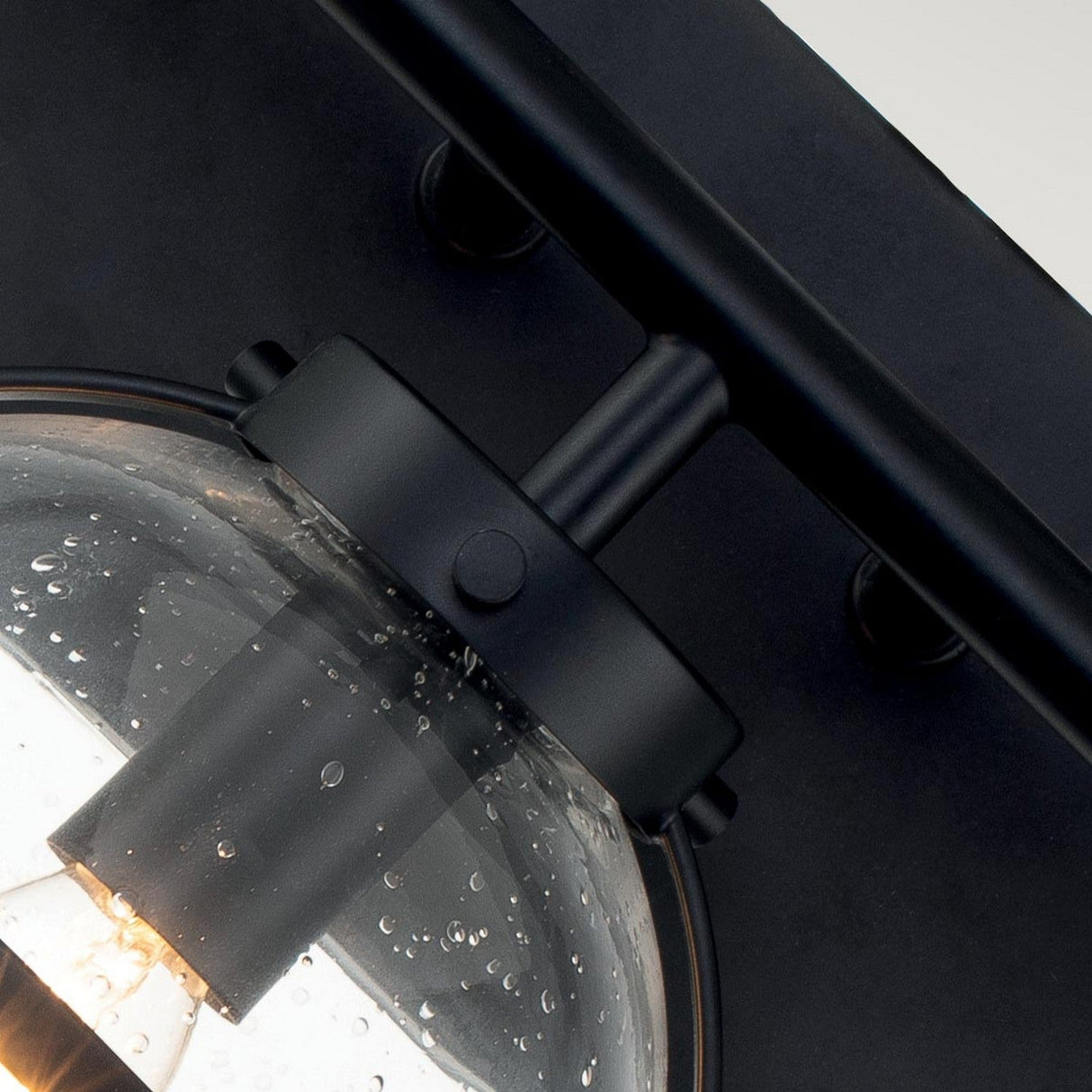 Close-up of the Hollis 3 Light Wall Light, featuring an Art Deco-inspired modern design with clear glass shades adorned with small bubbles and a sleek black finish. The illuminated bulb inside casts a warm glow, highlighting the elegant interplay between glass and metal.