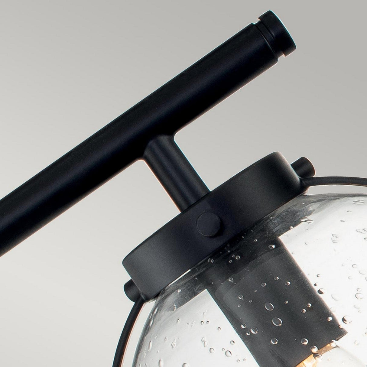 Close-up of the top portion of the Hollis 3 Light Wall Light - Black With Clear Glass, highlighting its black metal frame and a clear glass globe adorned with small bubbles. The fixture hangs gracefully against a neutral gray background, exuding elegance with its black finish.