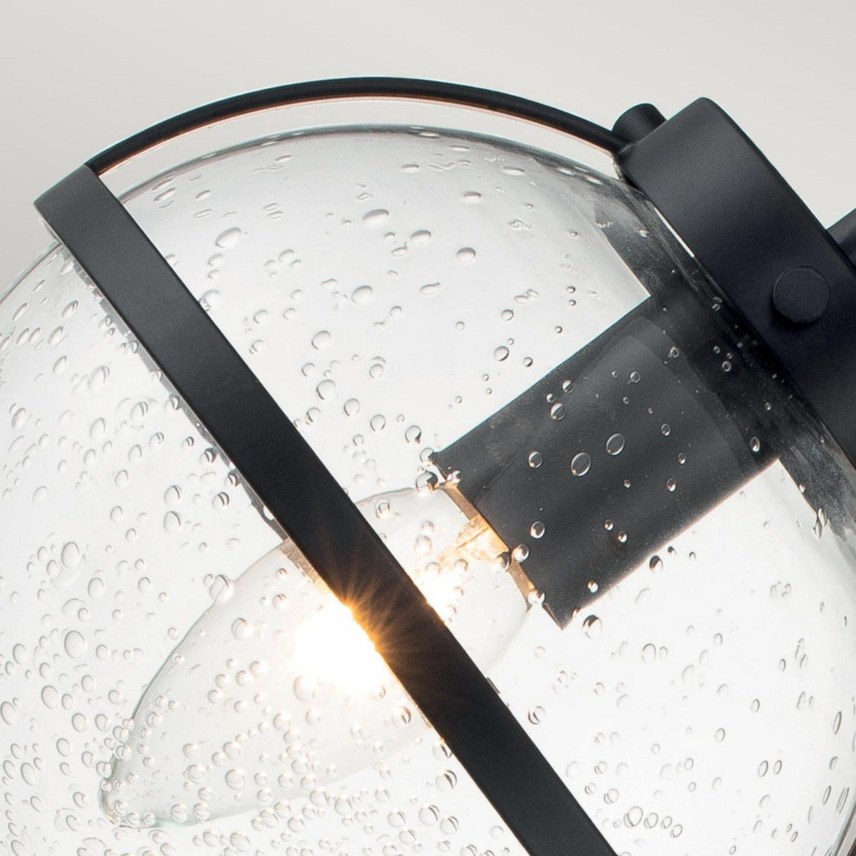 A close-up of the Hollis 3 Light Wall Light showcases its round clear glass lampshade featuring a water droplet texture reminiscent of Art Deco designs. A lit bulb inside is encased within a circular metal frame with a black finish, all set against a soft gray background.