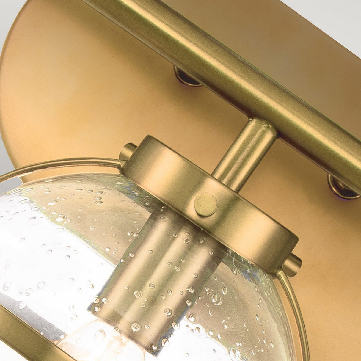 The close-up showcases a Hollis 3 Light Wall Light in Heritage Brass, featuring a clear glass globe. The glass includes small, decorative air bubbles that enhance the brushed metallic finish. This piece exudes modern elegance while seamlessly incorporating Art Deco inspiration, making it a sophisticated addition to any space.